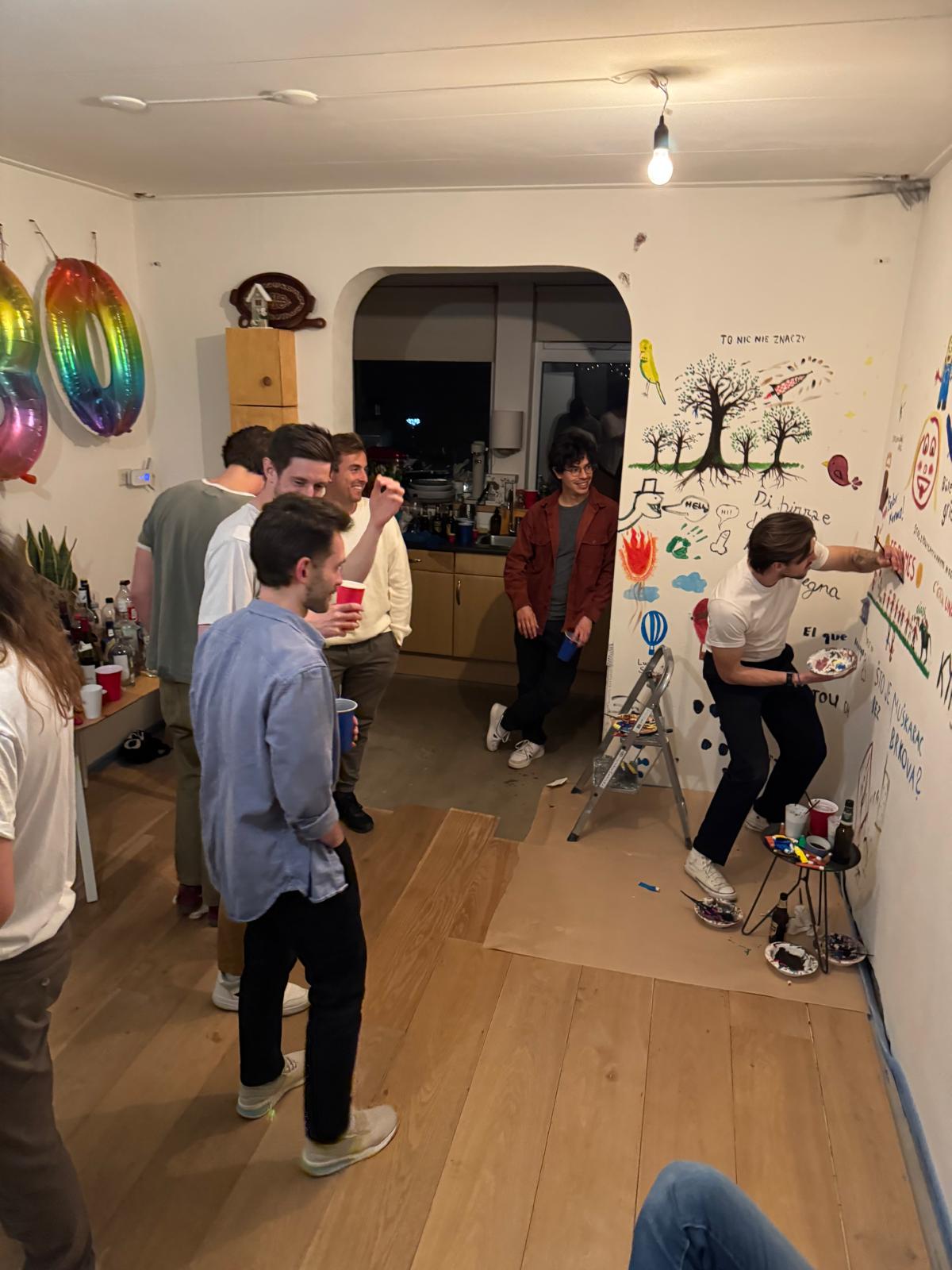 Greg painting on the wall at a house-warming party while friends watch